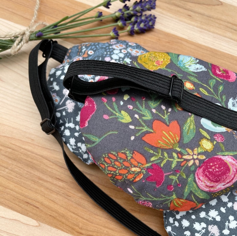 May include: A black sleep mask with an adjustable strap. The mask is made of fabric with a floral pattern in shades of pink, orange, yellow, and green on a dark gray background.