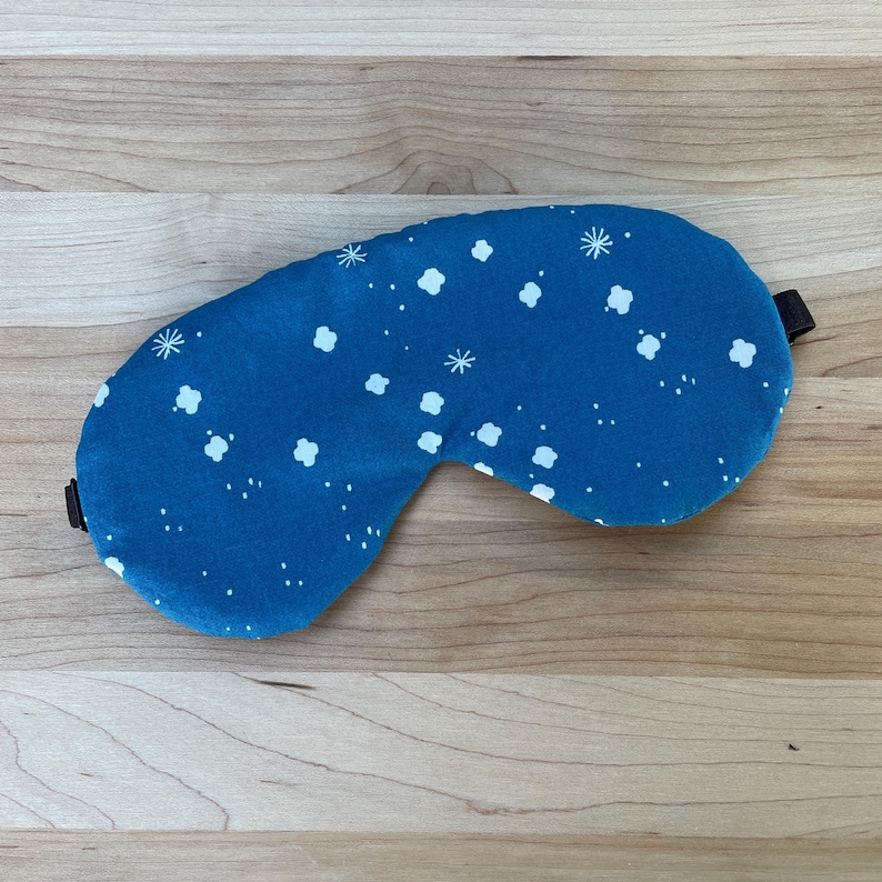 May include: A blue sleep mask with a white cloud and star pattern. The mask has a dark brown elastic strap and is designed to block out light. The sleep mask is made of soft fabric and is designed for comfort.