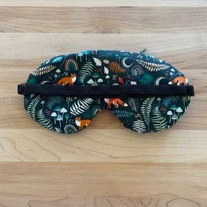 May include: A dark blue sleep mask with a woodland pattern featuring orange foxes, mushrooms, ferns, and rainbows. The mask has a black elastic strap for a comfortable fit. This eye mask is designed for relaxation and sleep.