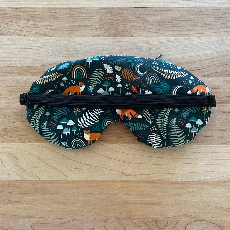 May include: A dark blue sleep mask with a woodland pattern featuring orange foxes, mushrooms, ferns, and rainbows. The mask has a black elastic strap for a comfortable fit. This eye mask is designed for relaxation and sleep.