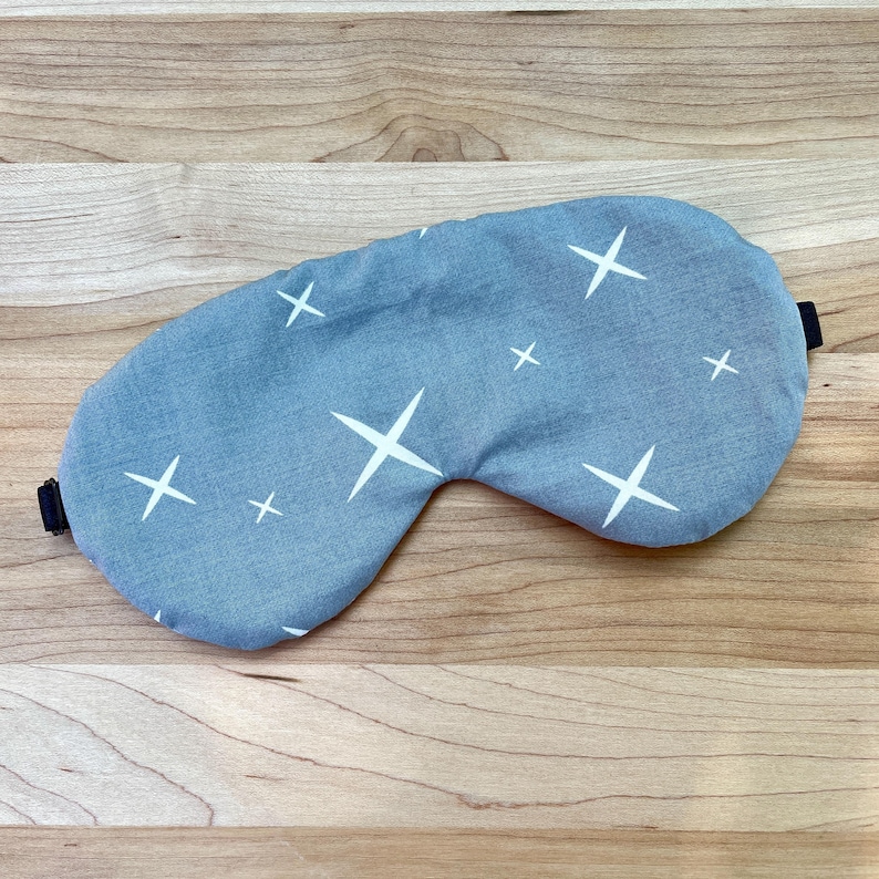 May include: A light blue sleep mask with a white star pattern. The mask has a black elastic band and is designed to block out light. The sleep mask is made of soft fabric and is designed for comfort.