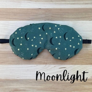 Puede incluir: Una m&aacute;scara para dormir de color verde azulado con un patr&oacute;n de lunas crecientes negras y peque&ntilde;as estrellas blancas. La m&aacute;scara tiene una banda el&aacute;stica negra y la palabra "Moonlight" en cursiva.