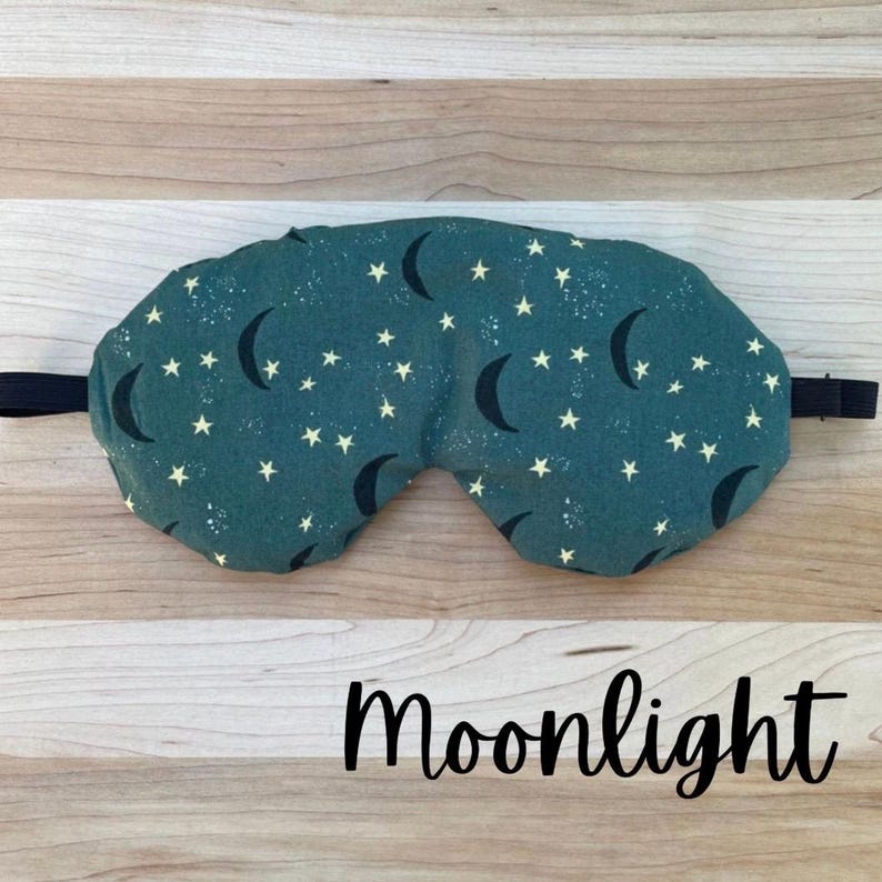 Puede incluir: Una m&aacute;scara para dormir de color verde azulado con un patr&oacute;n de lunas crecientes negras y peque&ntilde;as estrellas blancas. La m&aacute;scara tiene una banda el&aacute;stica negra y la palabra "Moonlight" en cursiva.