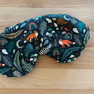 May include: A dark blue sleep mask with a whimsical forest pattern. The fabric features orange foxes, green ferns, white mushrooms, rainbows, and crescent moons. The mask is designed to block out light for better sleep.