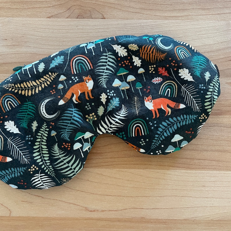 May include: A dark blue sleep mask with a whimsical forest pattern. The fabric features orange foxes, green ferns, white mushrooms, rainbows, and crescent moons. The mask is designed to block out light for better sleep.