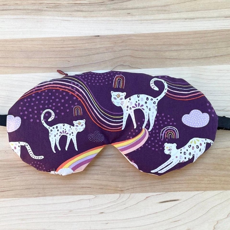 Puede incluir: Una m&aacute;scara para dormir morada con un dise&ntilde;o caprichoso de gatos blancos con manchas, arco&iacute;ris y nubes. La m&aacute;scara tiene una correa el&aacute;stica negra y una leng&uuml;eta roja. La m&aacute;scara para los ojos con tem&aacute;tica de gatos est&aacute; dise&ntilde;ada para la comodidad y el bloqueo de la luz.