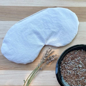 May include: A white linen eye mask with a curved shape, lying on a wooden surface. A sprig of dried lavender and a bowl of flax seeds are also visible.