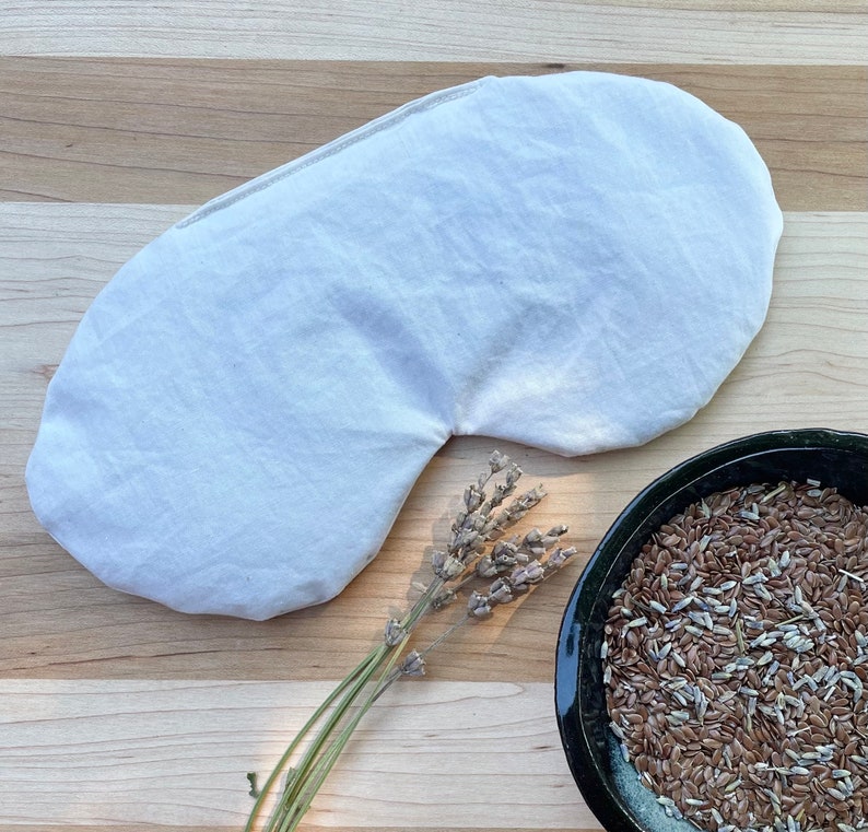 May include: A white linen eye mask with a curved shape, lying on a wooden surface. A sprig of dried lavender and a bowl of flax seeds are also visible.