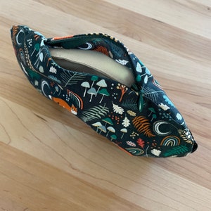 May include: A dark teal zippered pouch with a woodland pattern featuring mushrooms, ferns, foxes, and rainbows. The pouch is open, revealing a beige interior. The zipper is green.