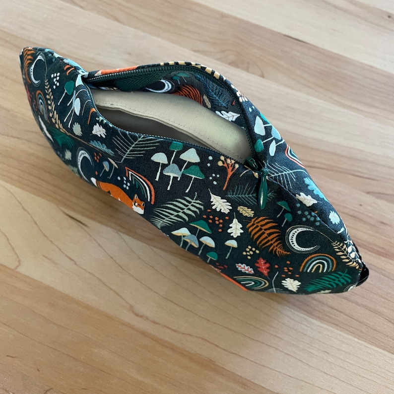 May include: A dark teal zippered pouch with a woodland pattern featuring mushrooms, ferns, foxes, and rainbows. The pouch is open, revealing a beige interior. The zipper is green.