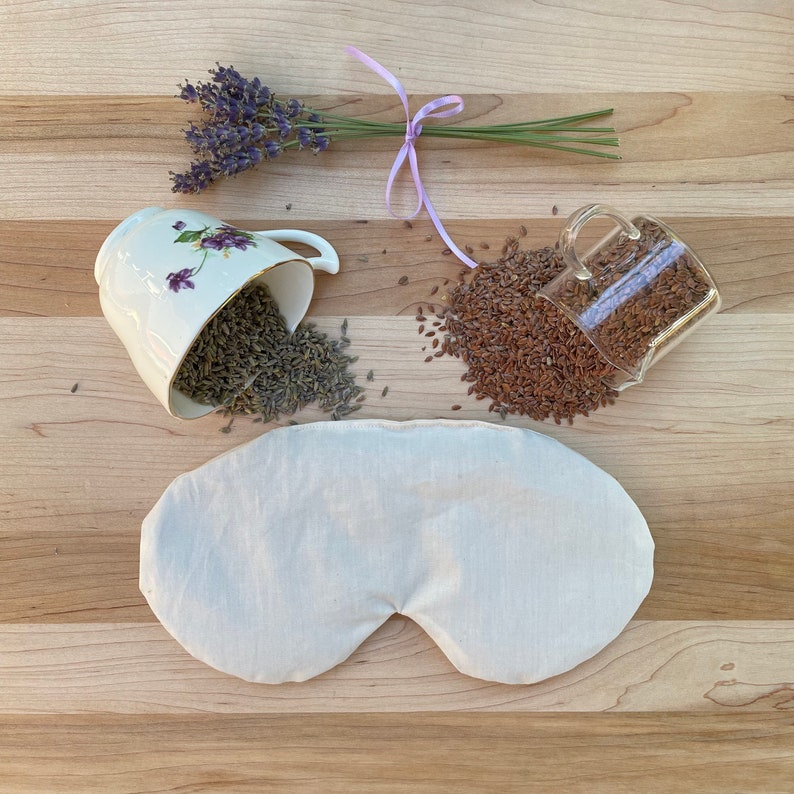 May include: A white eye mask filled with flax seeds and lavender for relaxation. The mask is on a wooden surface with a cup of lavender and a cup of flax seeds.