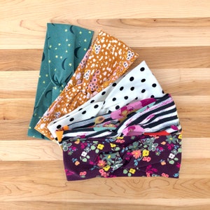 May include: A set of five fabric headbands in various patterns. The headbands are made of soft, stretchy fabric and feature a variety of colors and designs, including green with stars, orange with floral patterns, white with black polka dots, black and white stripes, and burgundy with floral patterns.