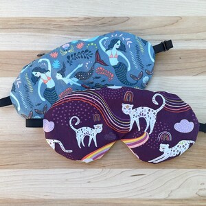 May include: Two sleep masks, one with a purple background and a white cat design, and the other with a blue background and a mermaid design.