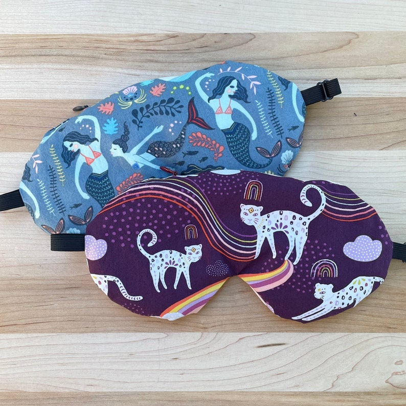 May include: Two sleep masks, one with a purple background and a white cat design, and the other with a blue background and a mermaid design.