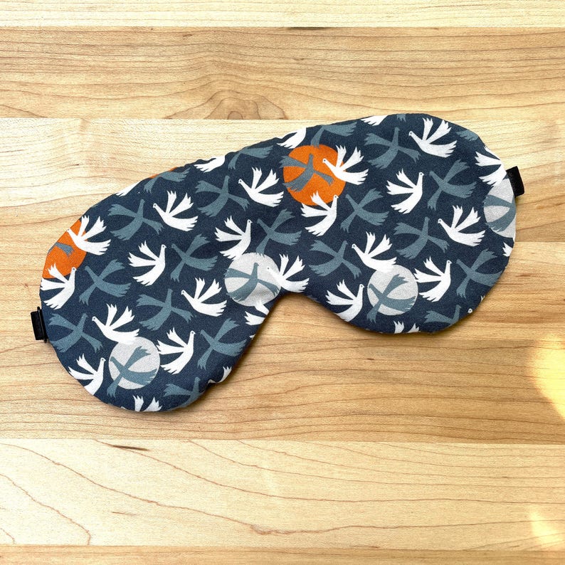 May include: A navy blue sleep mask with a repeating pattern of white birds and gray birds. The design includes orange and gray circles. The mask has a black elastic strap and is designed to block out light.
