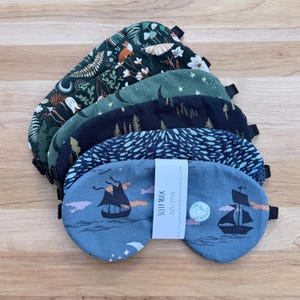 May include: A stack of five fabric sleep masks with various patterns including a blue mask with a ship and moon, a dark blue mask with small white shapes, a black mask with trees, a green mask with stars, and a dark green mask with floral motifs.
