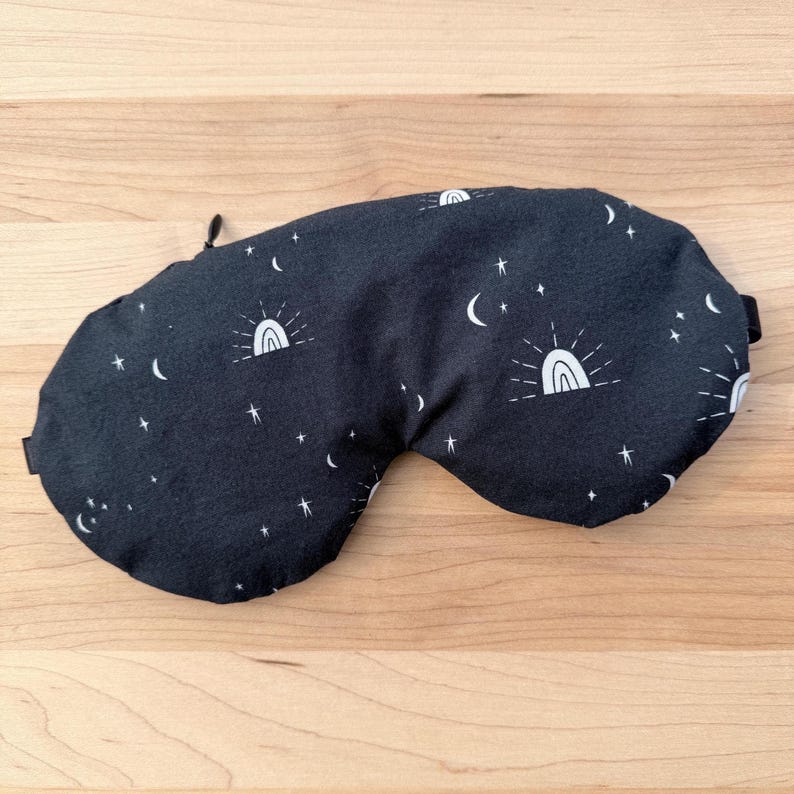 Puede incluir: M&aacute;scara para dormir negra con un patr&oacute;n celestial blanco de estrellas, lunas crecientes y arco&iacute;ris. La m&aacute;scara tiene una forma curva y una correa el&aacute;stica negra. Una peque&ntilde;a etiqueta negra est&aacute; adherida al lateral.
