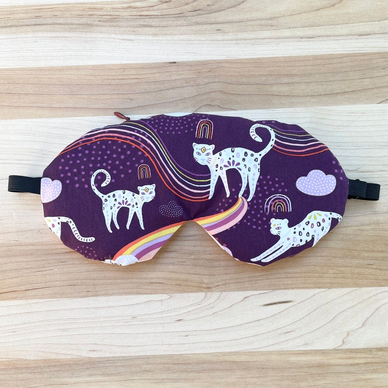 May include: Purple sleep mask with a white and black leopard print design. The mask features a rainbow and clouds.