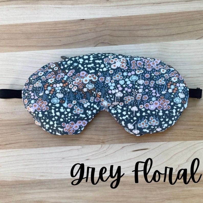 Puede incluir: Una m&aacute;scara para dormir floral gris con una banda el&aacute;stica negra. La m&aacute;scara presenta un patr&oacute;n de peque&ntilde;as flores en tonos rosa, blanco y azul. El texto "grey floral" est&aacute; escrito en cursiva debajo.