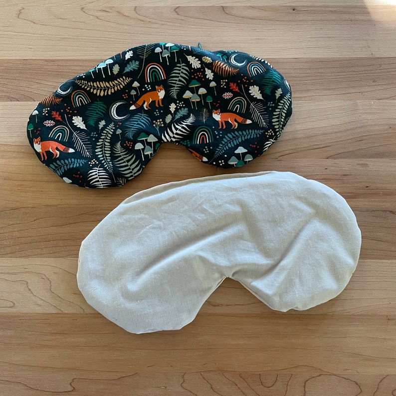May include: Two eye masks. One is patterned with foxes, mushrooms, rainbows, and foliage on a dark background. The other is a solid, light beige color. Both masks are designed to block out light.