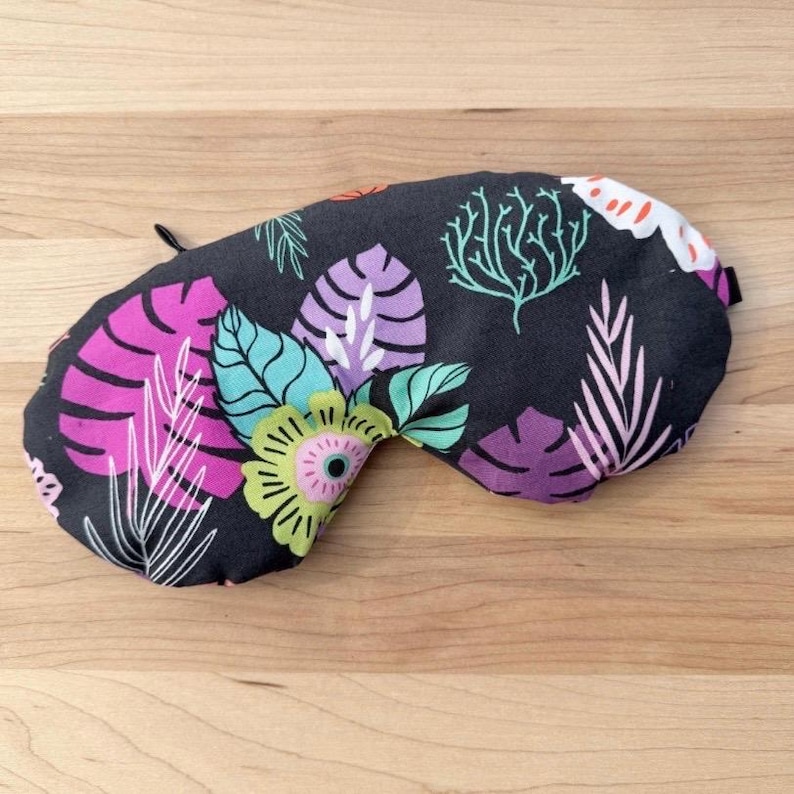 Puede incluir: Una m&aacute;scara para dormir negra con un vibrante estampado floral. La m&aacute;scara presenta grandes hojas y flores estilizadas en tonos de rosa, morado, turquesa y verde. El dise&ntilde;o est&aacute; sobre un fondo oscuro y est&aacute; dise&ntilde;ado para bloquear la luz.