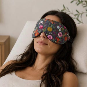 Könnte beinhalten: Eine Schlafmaske mit dunkelgrauem Hintergrund und einem lebendigen Blumenmuster in Rosa-, Gelb-, Orange- und Weißtönen. Die Maske hat eine konturierte Form zur Abdeckung der Augen und ist mit einem elastischen Band zur Befestigung versehen.