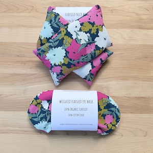 Pode incluir: Um conjunto de compressas para o pescoço de linhaça e uma máscara de olhos ponderada. Os produtos apresentam um padrão floral com flores rosa, brancas e amarelas sobre um fundo azul marinho. A máscara para os olhos é rotulada como "WEIGHTED FLAXSEED EYE MASK".