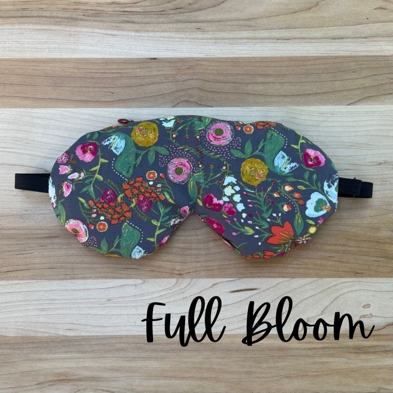 May include: A gray sleep mask with a floral pattern featuring pink, yellow, and green flowers. The text "Full Bloom" is printed below the mask.