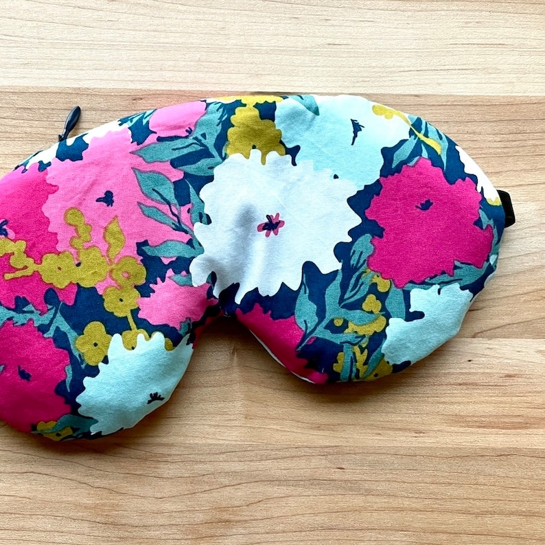 Puede incluir: Una m&aacute;scara para dormir floral con un patr&oacute;n de grandes flores rosas, blancas y amarillas y hojas verdes sobre un fondo azul marino. La m&aacute;scara tiene una correa el&aacute;stica negra y un peque&ntilde;o lazo en el lateral.
