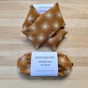 Pode incluir: Um conjunto de compressas para o pescoço e uma máscara de olhos com sementes de linhaça. Os produtos são feitos de tecido castanho com um padrão de sol branco. Os rótulos dizem "100% Organic Flaxseed" e "100% Cotton Cover".