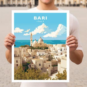 Pode incluir: Um poster com uma ilustração estilizada de Bari, Itália, com o texto "BARI ITALY" no topo. A obra de arte retrata edifícios brancos, uma igreja com cúpulas, uma alta torre sineira e um céu azul com nuvens. O poster é segurado por uma pessoa.