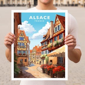 May include: A vintage travel poster featuring a colorful illustration of a street scene in Alsace, France. The poster has the text "Alsace, France" in a bold, white font.