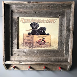 May include: A rustic wooden wall-mounted gun rack with a framed vintage Remington-UMC advertisement featuring two black Labrador puppies. The rack has four shotgun shell hooks.
