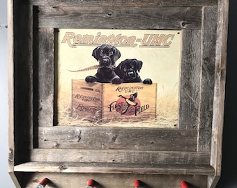 May include: A rustic wooden wall-mounted gun rack with a framed vintage Remington-UMC advertisement featuring two black Labrador puppies. The rack has four shotgun shell hooks.