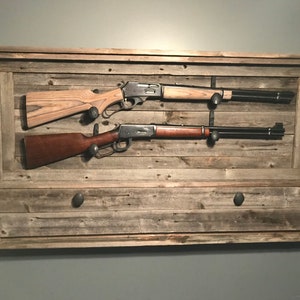 May include: A rustic wooden wall mount with a drawer, designed to hold two rifles. The rifles are secured with hooks and are displayed on a wood plank background.