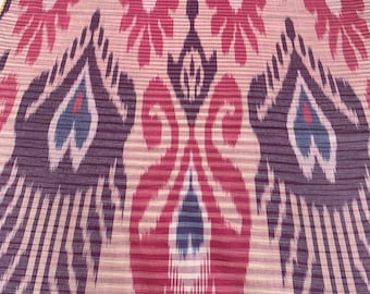 May include: A close-up of a fabric with a pink, purple, and blue ikat pattern. The pattern features a repeating design of stylized flowers and leaves.