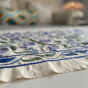 May include: A white linen table runner with blue and green embroidered floral designs. The runner has a delicate, hand-stitched look.