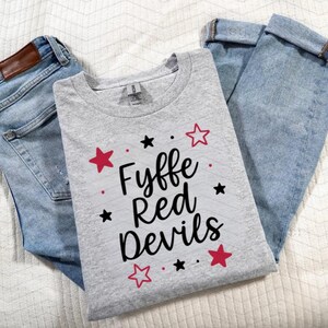 Puede incluir: Una camiseta gris claro con el texto "Fyffe Red Devils" en escritura negra, acentuada con estrellas rojas y negras. La camiseta se muestra con vaqueros de mezclilla azul claro.
