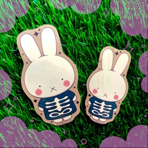 May include: Two white bunny stickers with black eyes and pink cheeks. Both bunnies are wearing a blue shirt with a white skeleton design. The stickers are on a green grass background. The text '@UBEPIG' is on the bottom of each sticker.