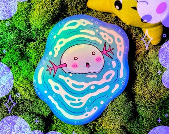 May include: A holographic sticker featuring a cartoon axolotl with a pink body and blue eyes, swimming in a swirling blue and white pattern. The sticker has a white border and the text "U.B.E.P.G." in a black font.