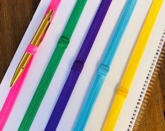 May include: A white notebook with six colorful elastic bands attached to the spine. The bands are pink, green, purple, blue, yellow, and orange. A gold pen is placed on top of the notebook.