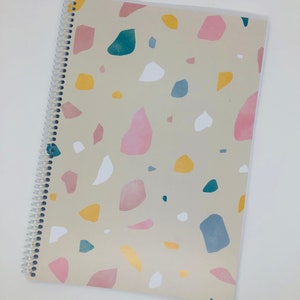 May include: A spiral bound notebook with a beige cover featuring a terrazzo pattern in shades of pink, blue, yellow, and white.