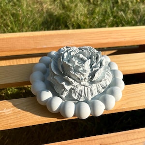 May include: A light gray decorative object featuring a central sculpted flower with layered petals, surrounded by a circular border of uniform spherical beads. The entire piece has a matte finish and a consistent monochromatic light gray color.