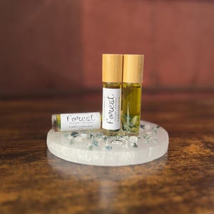 May include: Three essential oil roller bottles with bamboo caps and the word "Forest" on the labels. The bottles sit on a white, round tray with small, blue-green stones. The background is a warm brown.