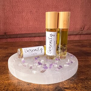 May include: Three essential oil roller bottles with bamboo lids and labels that read "Serenity". The bottles sit on a white, round stone base with scattered purple crystals. The background is a solid, reddish-brown color.