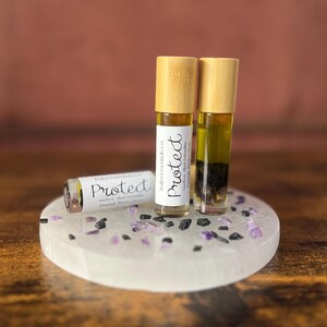 May include: Three essential oil roller bottles with bamboo lids labeled "Protect" on a white stone base with purple and black crystals. The bottles contain a clear liquid with dark inclusions. The background is a blurred brown and pink.
