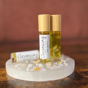 May include: Three glass roll-on bottles with wooden caps, labeled "Cleansing" and "Orange Clear Quartz High Vibration". The bottles are filled with a yellow liquid and sit on a white, round, crystal-covered surface. The background is a blurred brown.