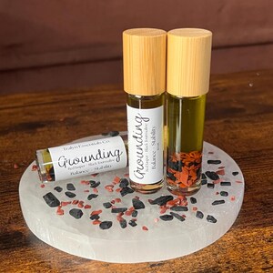 May include: Three essential oil roller bottles with wooden caps labeled "Grounding" on a white stone tray. The bottles contain red jasper and black tourmaline. The tray is decorated with black and red stones.