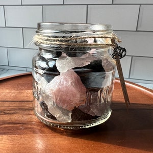 May include: A clear glass jar filled with various crystals, including black, clear, and pink stones. The jar is wrapped with twine and has a decorative charm attached. The jar sits on a wooden tray against a white tiled background.