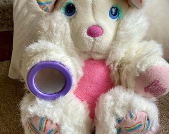 May include: White plush toy with a pink heart, rainbow accents, and purple eyes. The toy has a purple ring on its paw and a purple brush on the floor.
